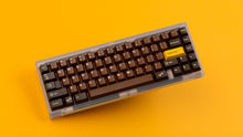 Load image into Gallery viewer, GMK CYL Chocolatier keycaps on clear transparent 65% keyboard against orange background, full angled view with golden Golden Ticket key