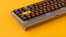 Load image into Gallery viewer, GMK CYL Chocolatier keycaps on clear transparent 65% keyboard against orange background, close-up of right side showing golden Golden Ticket key