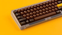 Load image into Gallery viewer, GMK CYL Chocolatier keycaps on clear transparent 65% keyboard against orange background, close-up of left side alternate angle