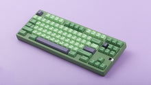 Load image into Gallery viewer, GMK CYL Zooted keycaps on green 1800 compact keyboard, front-angled wide shot showing full layout with purple spacebar and navigation cluster on lavender surface